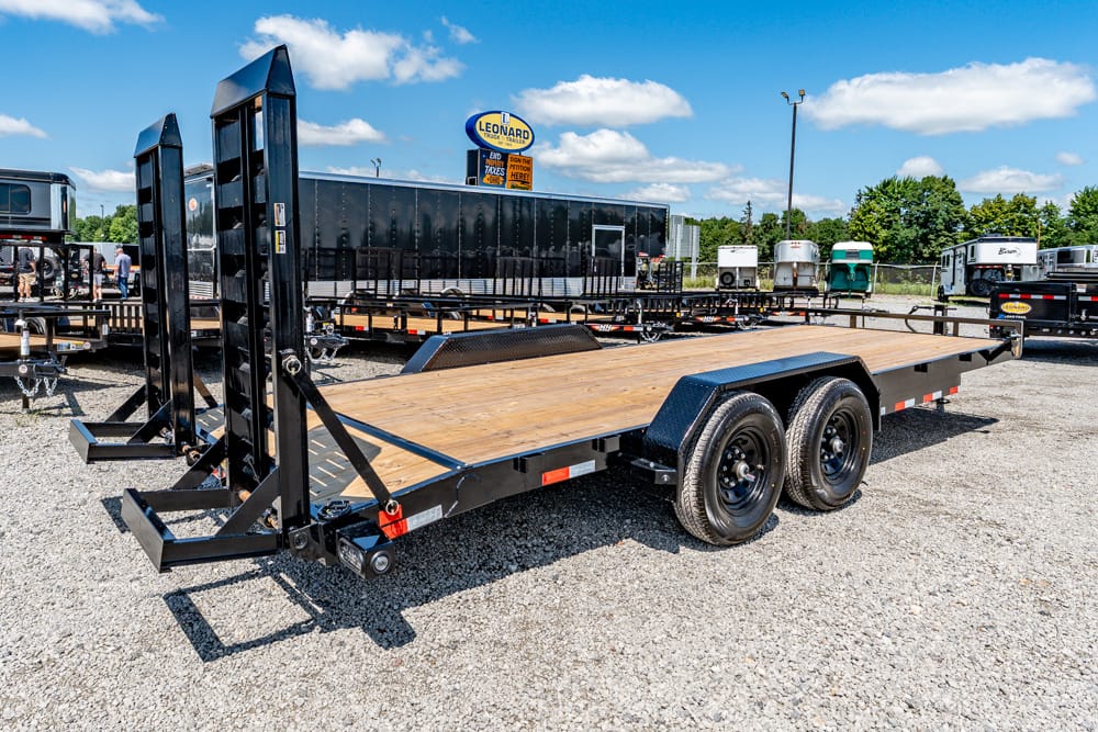 Black heavy-duty flatbed trailer with wooden deck and dual rear ramps in a gravel lot.