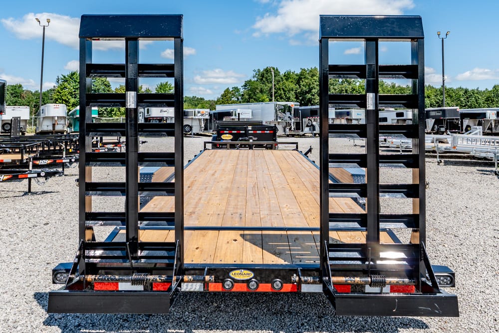 Black steel trailer with two tall loading ramps and a wooden deck in a gravel lot.