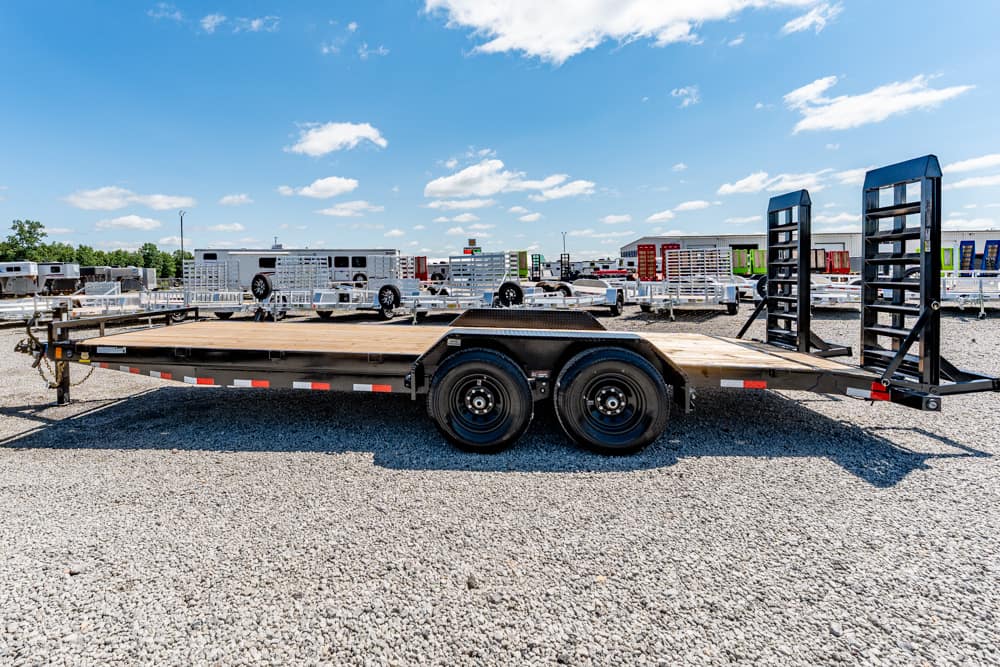 Flatbed trailer with dual rear axles parked on a gravel lot, with other trailers in the background and loading ramps at the front and rear.