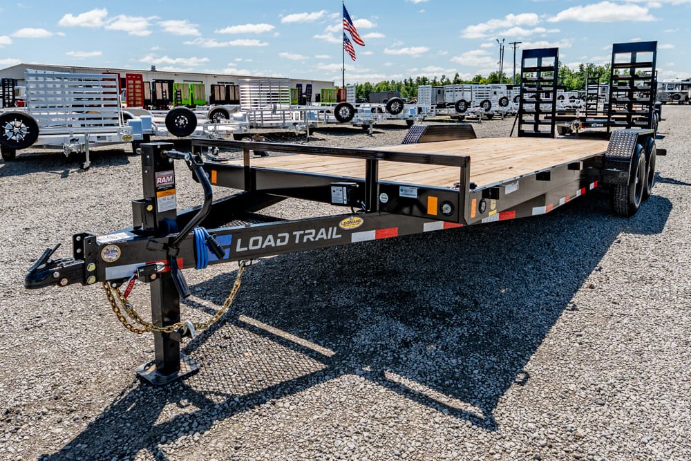 Flatbed trailer with a wooden deck and front hitch parked on a gravel lot, ramps at the rear.