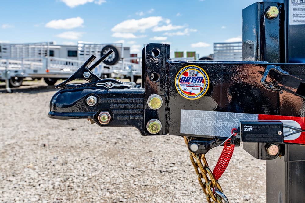 Close-up of a black trailer hitch with safety pins, bolts, and a red safety chain, set in a gravel lot with other trailers in the background