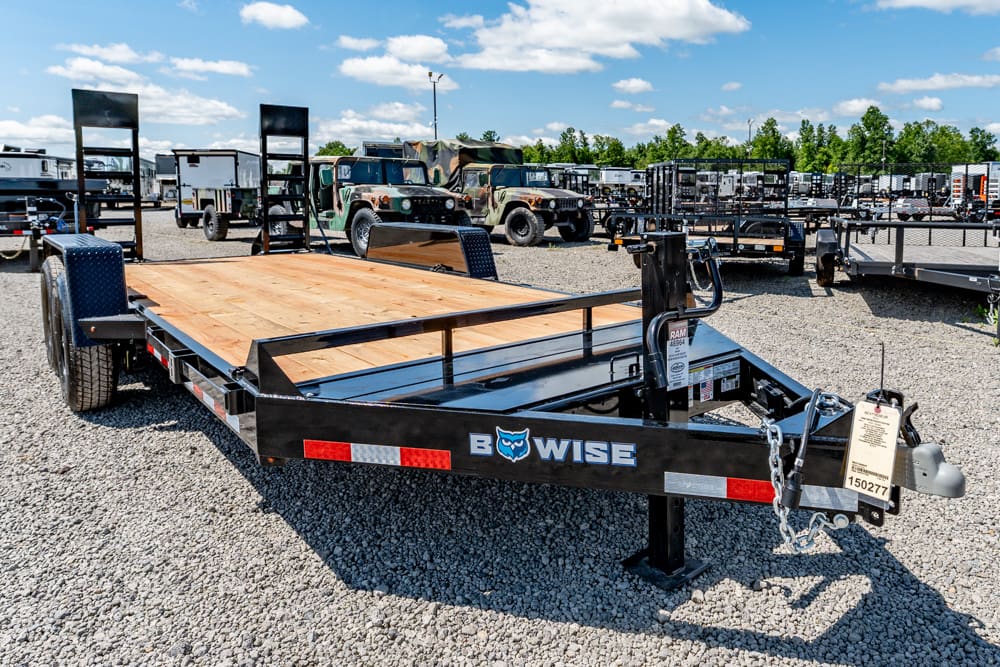 Black flatbed trailer with a wooden deck in a gravel lot, brand name BWise visible on the side and other trailers and trucks in the background.