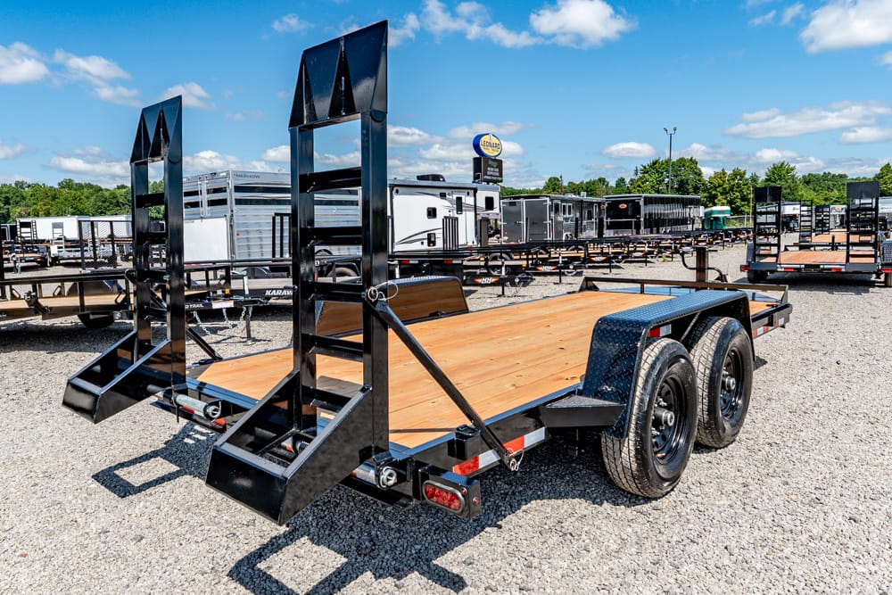 Flatbed trailer with fold-down ramps and dual axles parked on a gravel lot; other trailers and RVs visible in the background.