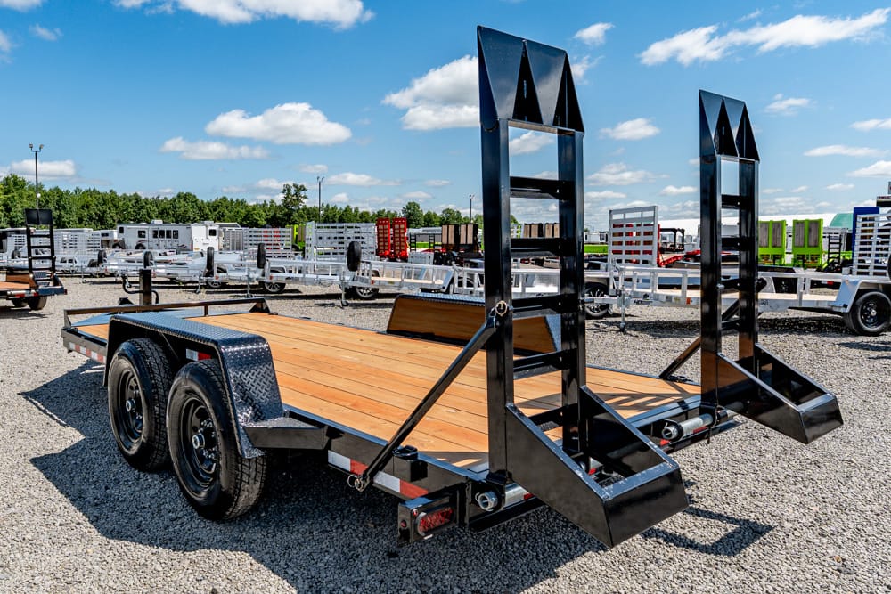 Black dual-axle flatbed trailer with a wooden deck and tall foldable ramps raised at the rear, parked on a gravel lot with other trailers nearby.