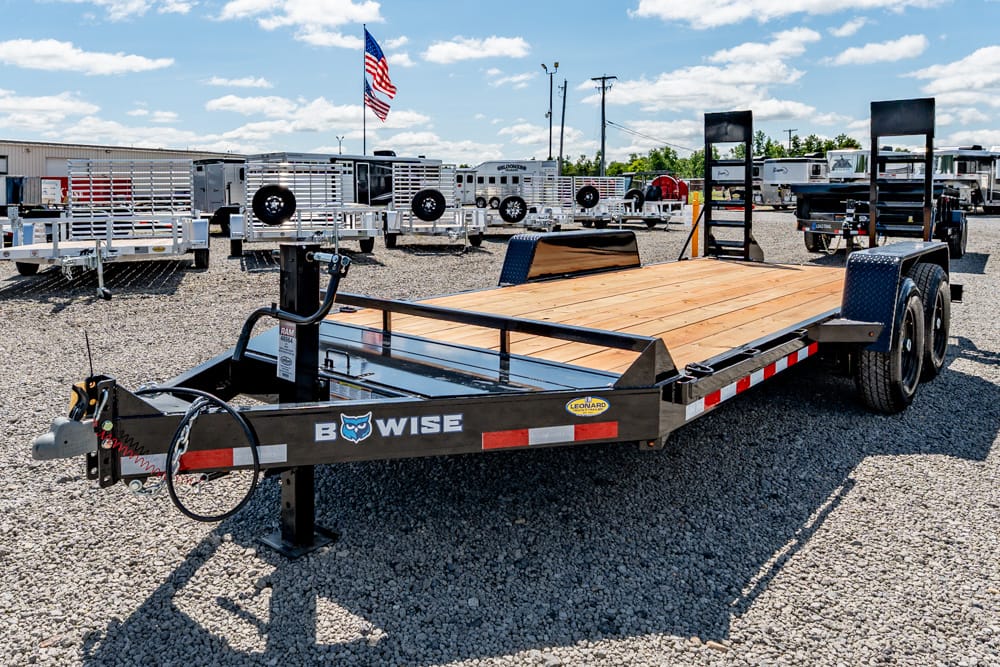 Dual-axle flatbed trailer with a wooden deck and ramps sits in a gravel trailer yard.