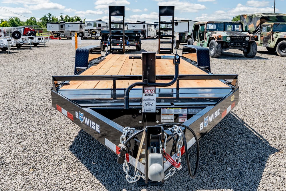 Front view of a BWISE flatbed trailer with a wooden deck and safety chains, on a gravel lot with other trailers in the background
