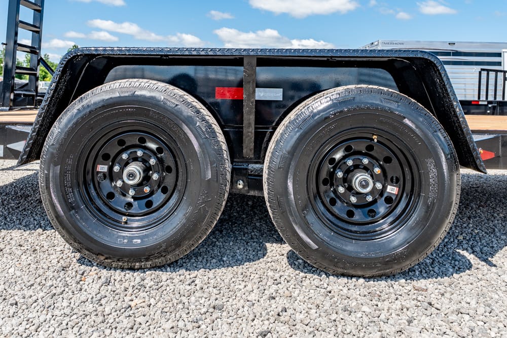 Tandem trailer wheels with black rims on a gravel surface, side view of the axle and tire treads under a black trailer fender