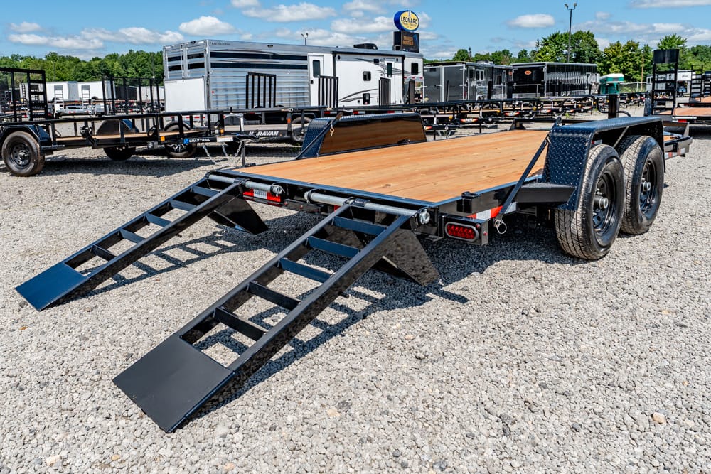 Dual-axle trailer with a wooden deck and two fold-down loading ramps extended, parked on a gravel trailer lot.
