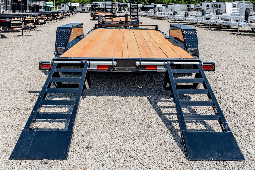 Flatbed trailer with wooden deck and lowered black loading ramps on a gravel lot, surrounded by other trailers.