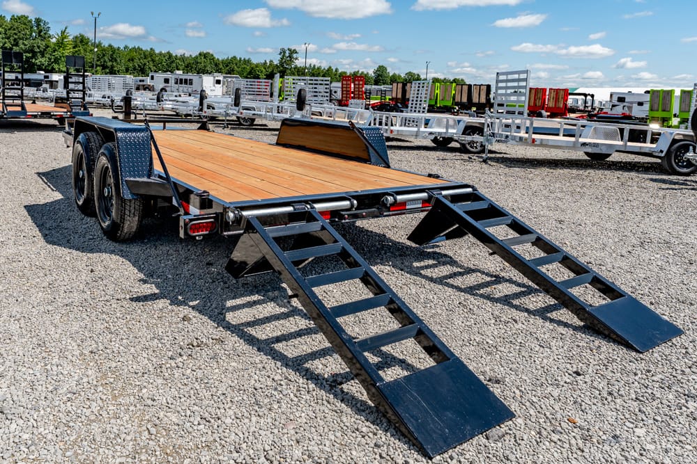 Flatbed trailer with a wooden deck and two fold-down ramps on a gravel lot, surrounded by other trailers in the background.