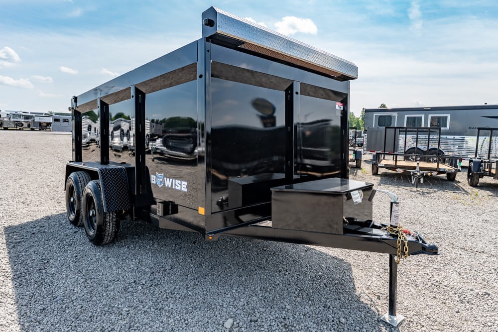 Black, dual-axle dump trailer on a gravel lot, front toolbox and hitch visible with a 'Wise' logo on the side, ready for use