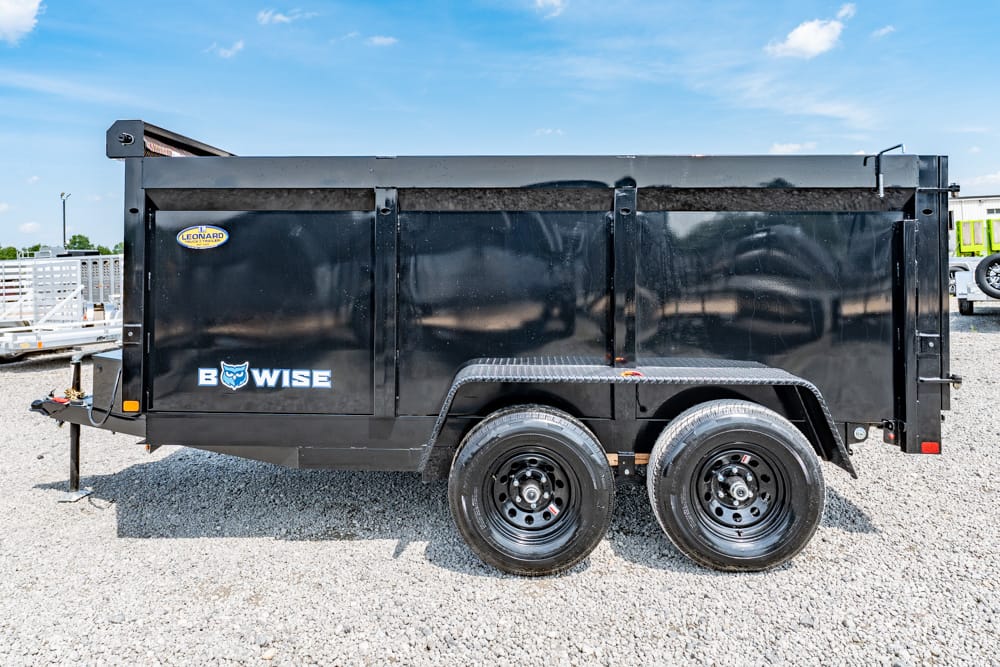 Black dual-axle dump trailer with a raised bed on a gravel lot, branded 'B WISE' and a small yellow decal on the side