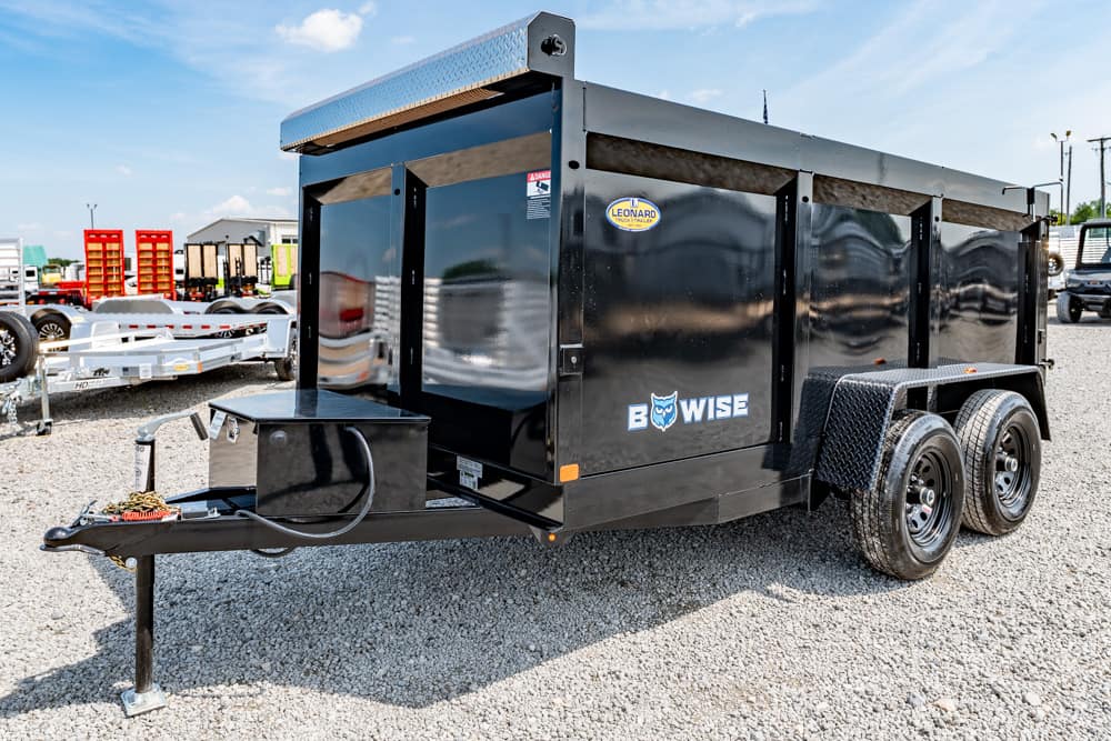Black Be Wise dump trailer with dual axles on a gravel lot, featuring a diamond-plate toolbox area and hitch at the front.