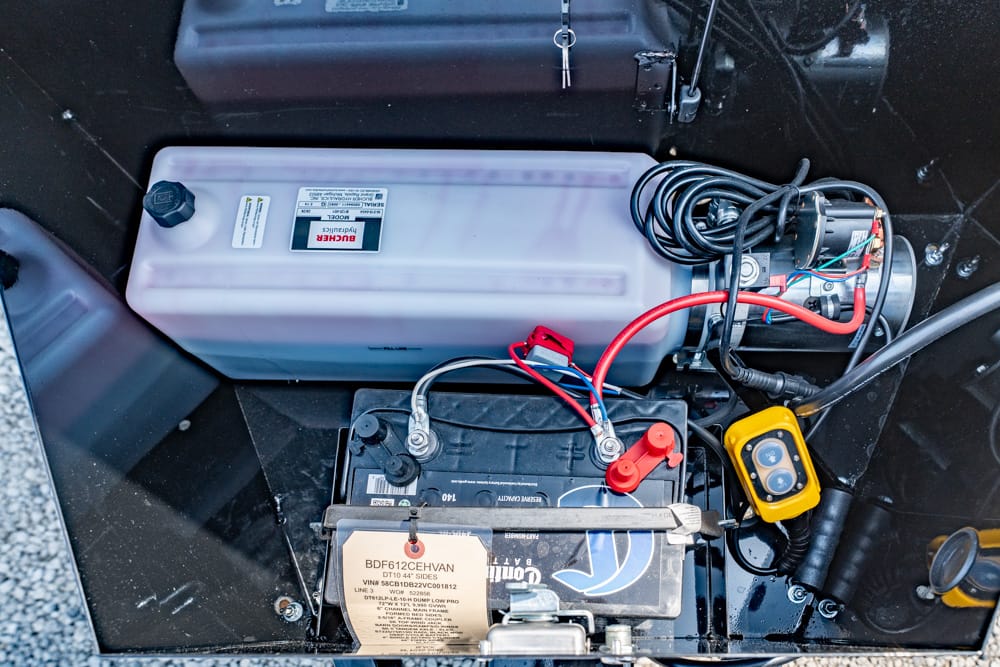 Open battery compartment showing a 12V battery with red and black cables connected, plus a yellow handheld meter nearby. A translucent white reservoir sits above.