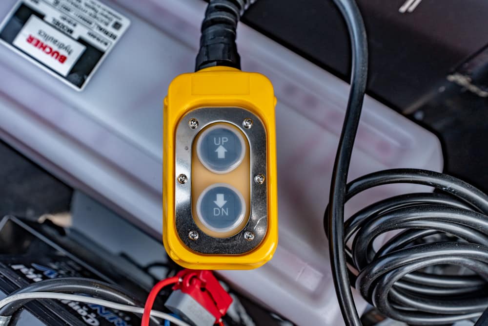 Yellow industrial control pendant with two round buttons labeled UP and DN (up/down) on a metal plate, connected by a cable to equipment.