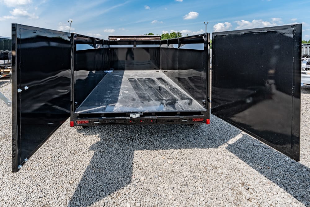 Black dump trailer with rear doors wide open, exposing an empty metal bed on a gravel lot under a blue sky.