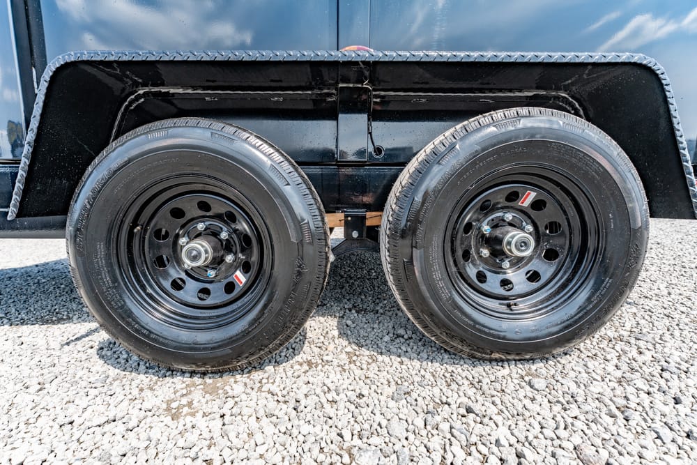 Dual-axle trailer wheels: two black rims and tires on a gravel surface beneath a black fender bottom edge.