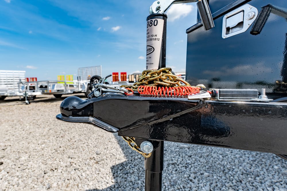 Close-up of a black trailer hitch with safety chains and a red coiled air hose on a gravel lot below a blue sky.