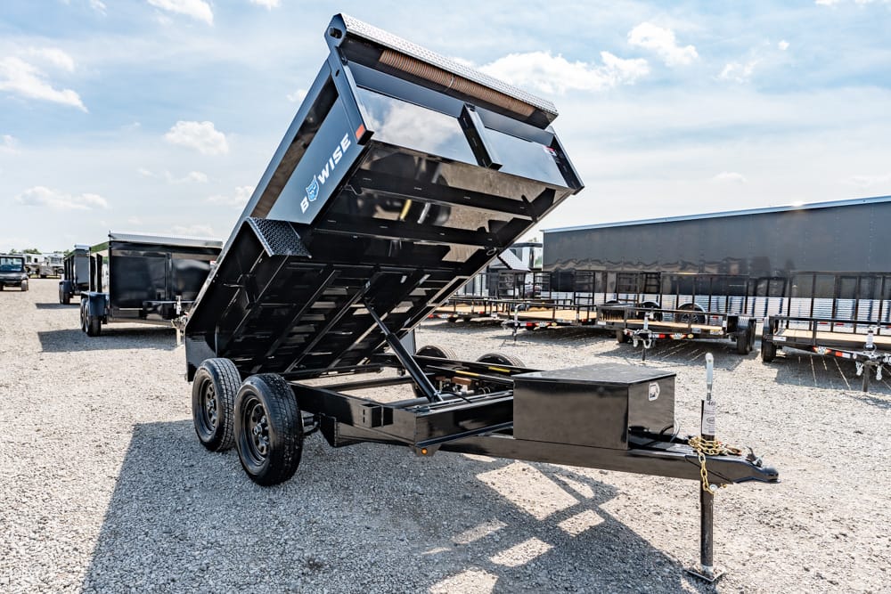 Tilt-bed dump trailer with hydraulic bed raised on a gravel lot, showing a dual-axle trailer connected to a hitch and a forward jack stand.