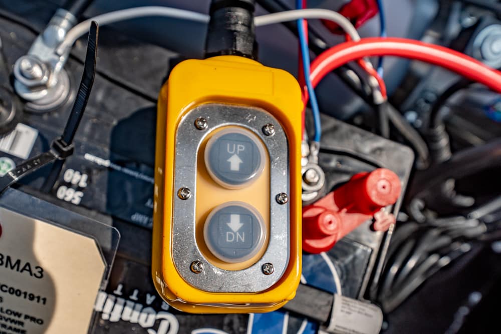 Close-up of a yellow two-button control with UP and DN arrows mounted on a car battery, surrounded by red and blue wires and battery terminals.