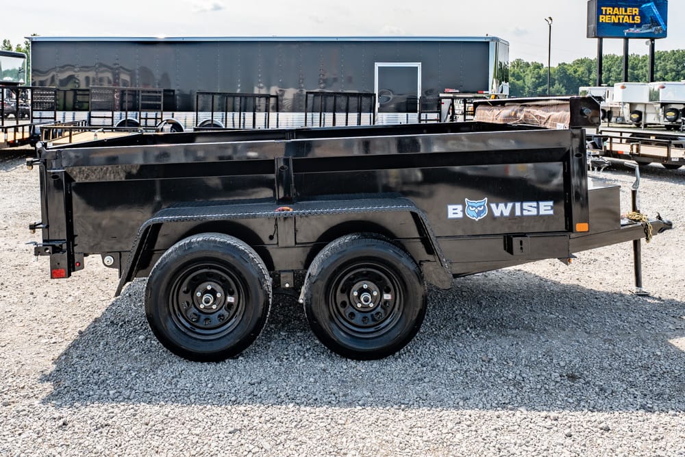 Black dual-axle dump trailer on a gravel lot with a 'B WISE' logo on the side and trailers in the background.