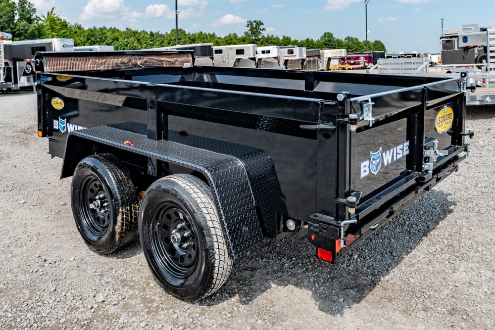 Black tandem-axle dump trailer with diamond-plate fenders in a gravel trailer yard, branding visible on sides.