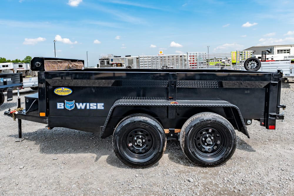 Black dual-axle utility trailer on a gravel lot, branded BOWISE with a metal toolbox and ramp sides visible in the background.