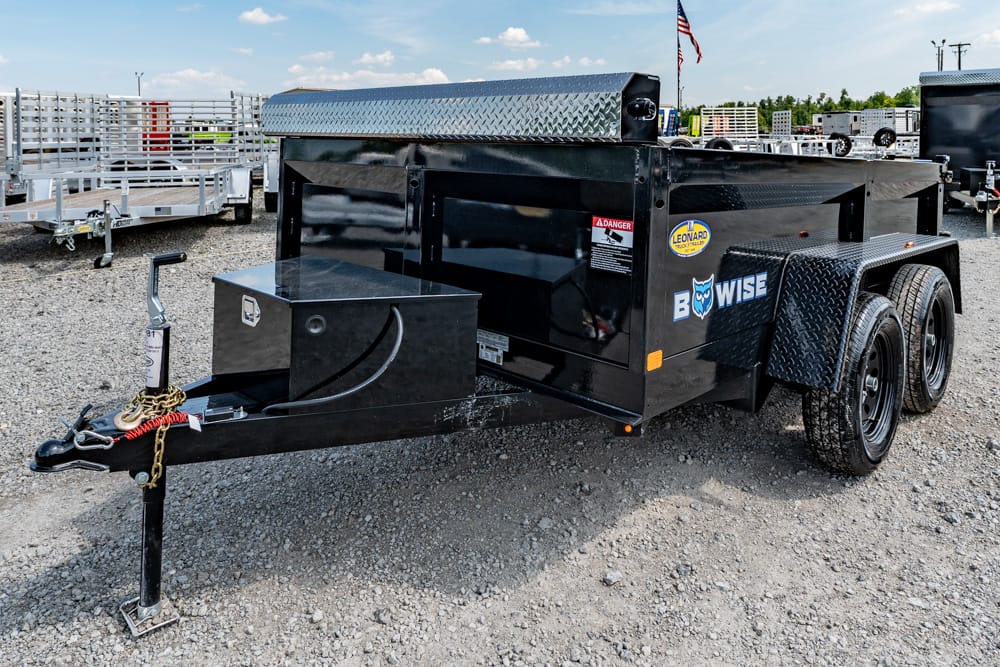Black dual-axle utility trailer with a diamond-plate toolbox at the front, parked on a gravel lot.