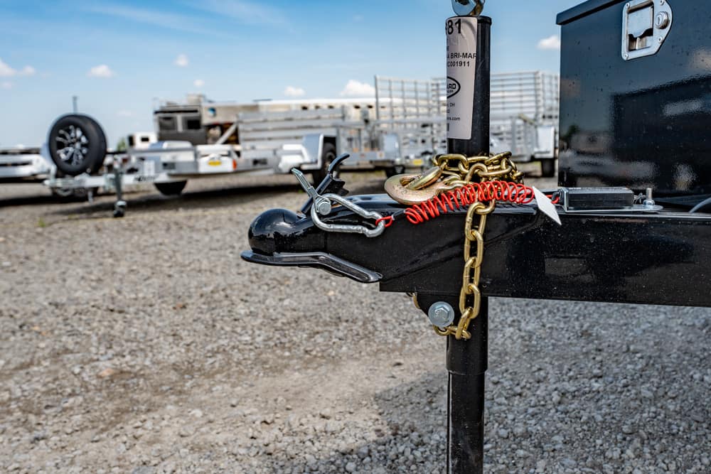Close-up of a black trailer hitch with a gold chain and red coiled cable secured across it in a gravel lot, with trailers in the background.