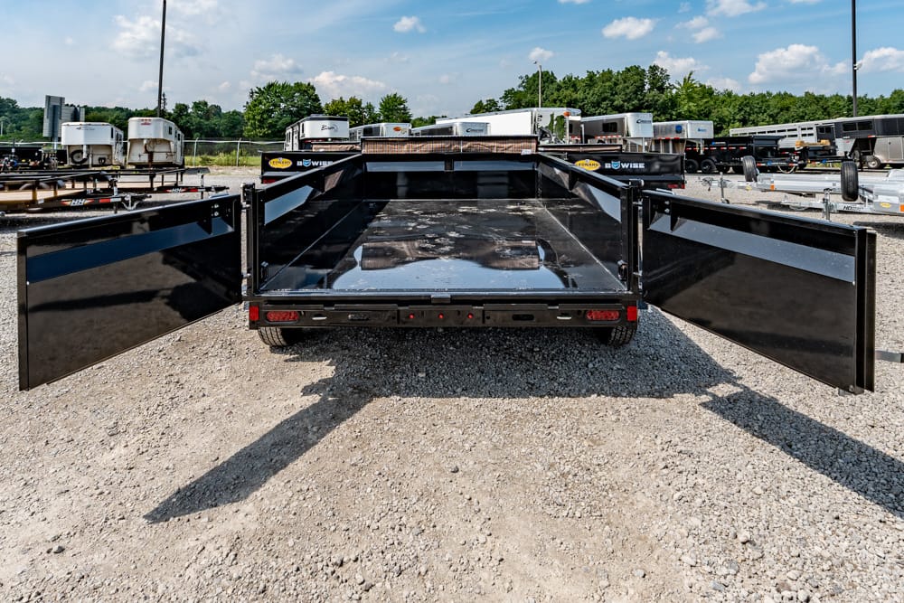 Back view of a black flatbed trailer with its rear doors wide open, parked on a gravel lot with other trailers in the background.