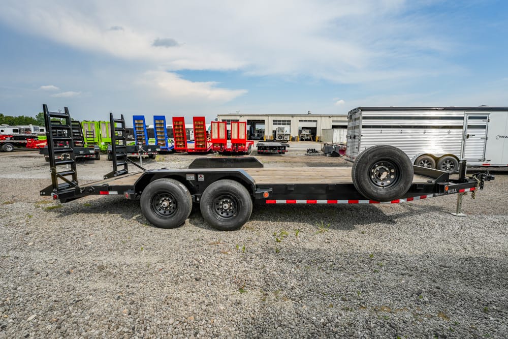 Flatbed trailer with dual axles and a spare tire mounted on the front, parked on a gravel lot with colorful equipment in the background.