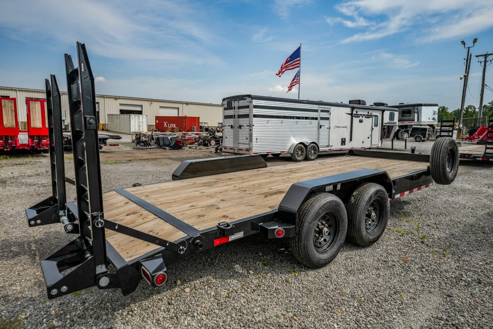 Three-axle car hauler trailer with a wooden deck and attached ramps in a gravel lot, ready to haul vehicles.
