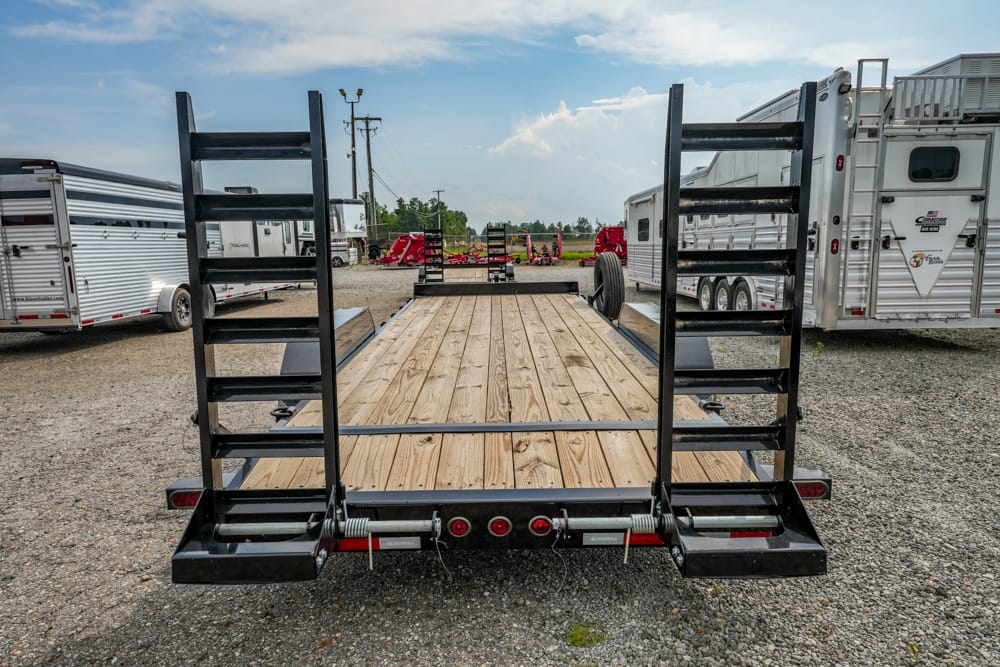Flatbed trailer with a wooden deck and tall black ramps on both sides, parked in a gravel lot with other horse trailers nearby