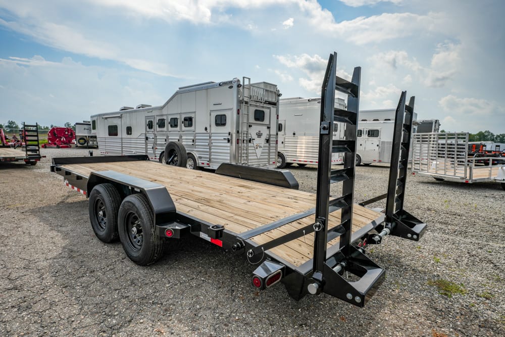 Black dual-axle flatbed trailer with wooden deck and dual fold-down ramps raised at the rear, parked on a gravel lot near horse trailers.