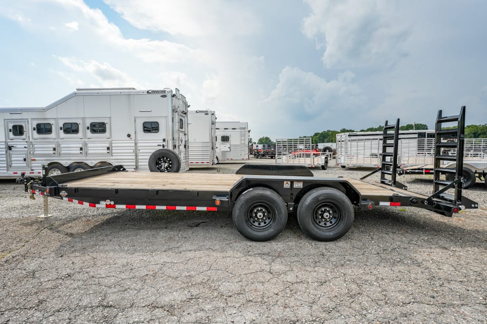 Flatbed trailer with dual axles and fold-up loading ramps in a gravel lot, with horse trailers in the background.