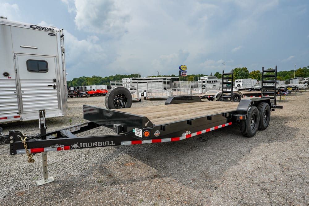 Black Iron Bull flatbed trailer with wooden deck in a gravel lot, two axles, spare tire, and a silver horse trailer nearby.