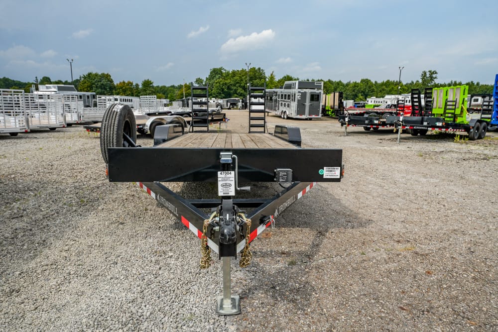 Front view of a black flatbed trailer on a gravel lot, with a spare tire, hitch, and other trailers in the background.