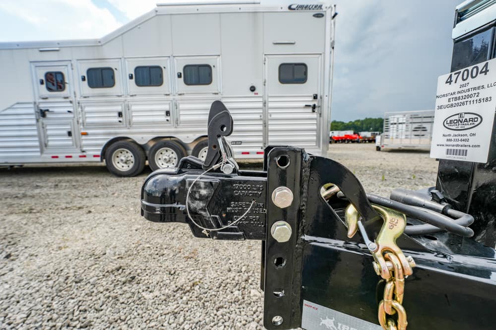 Close-up of a trailer hitch with a safety chain and jack on a black trailer, gravel lot with a silver horse trailer in the background.