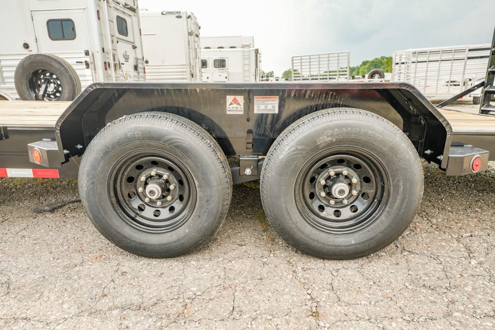 Dual-axle flatbed trailer with black fenders and heavy-duty tires on a gravel lot, with trucks in the background.