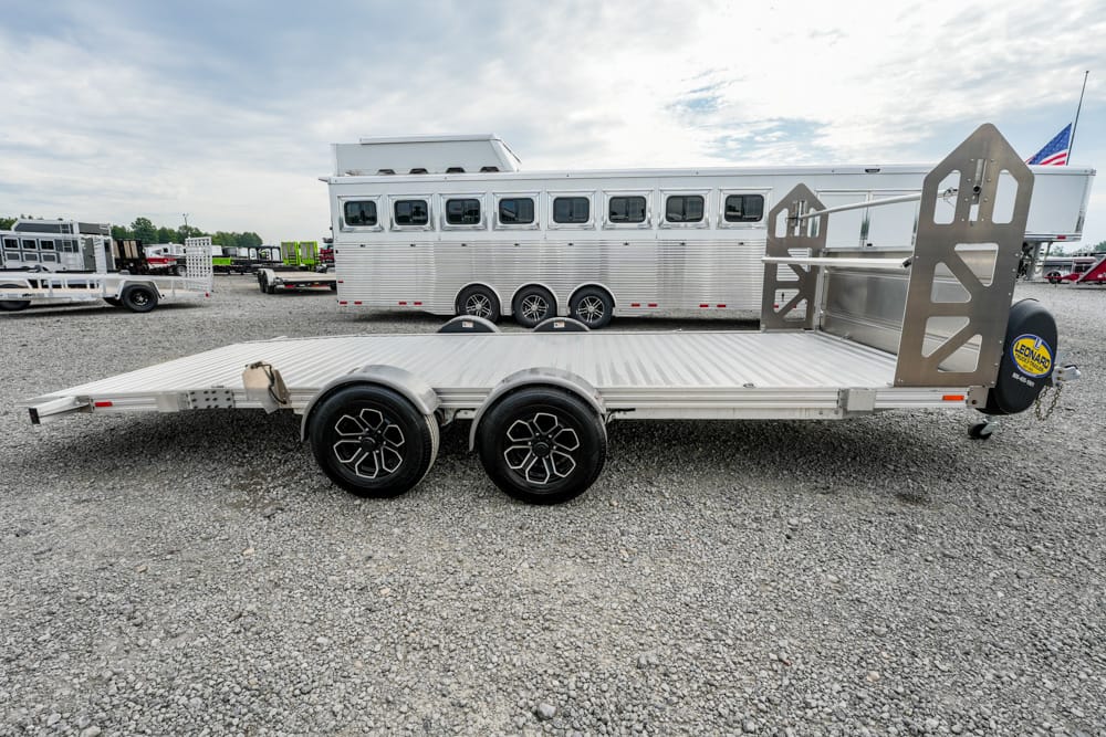 Aluminum two-axle flatbed trailer with a front spare tire and rear loading ramps, parked on a gravel lot with a large horse trailer in the background.