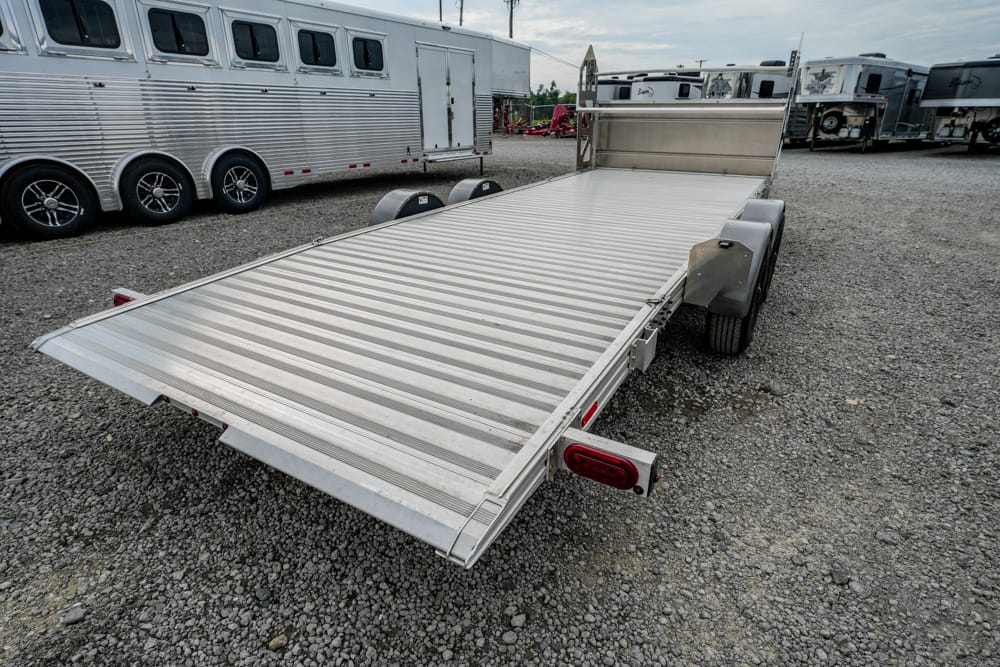 Aluminum flatbed trailer with a ramp, parked on a gravel lot among other trailers.