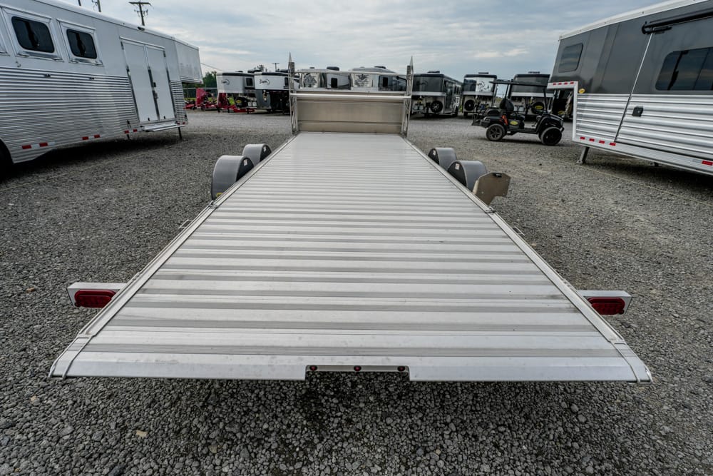 Aluminum flatbed trailer in a dealer lot with other trailers on a gravel surface under a cloudy sky