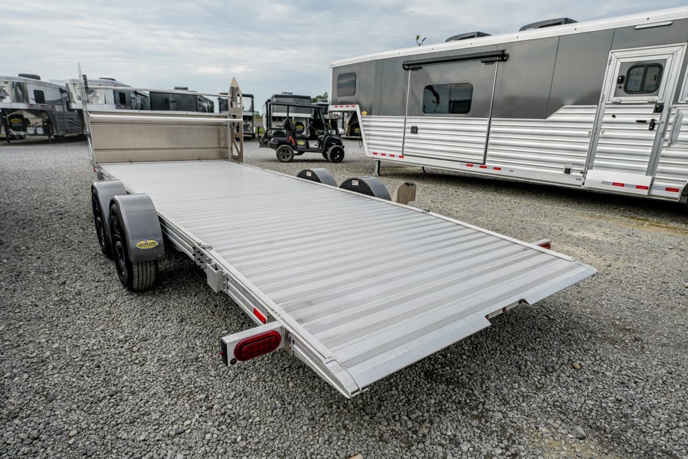 Aluminum flatbed trailer with tandem axles on a gravel lot, RVs and trailers in the background.