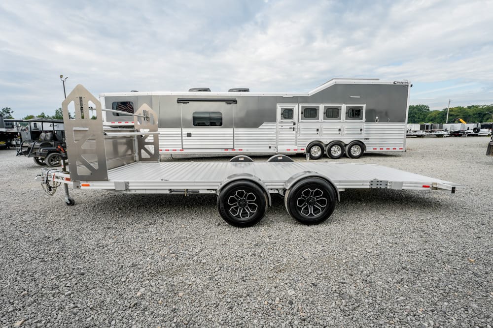 Aluminum flatbed car hauler trailer with dual axles and a rear ramp, parked on a gravel lot beside a large horse trailer.