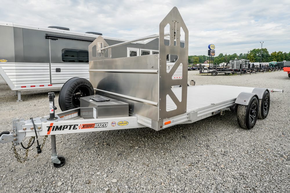 Aluminum car trailer with dual axles, spare tire, and a high side ramp, parked on a gravel lot.
