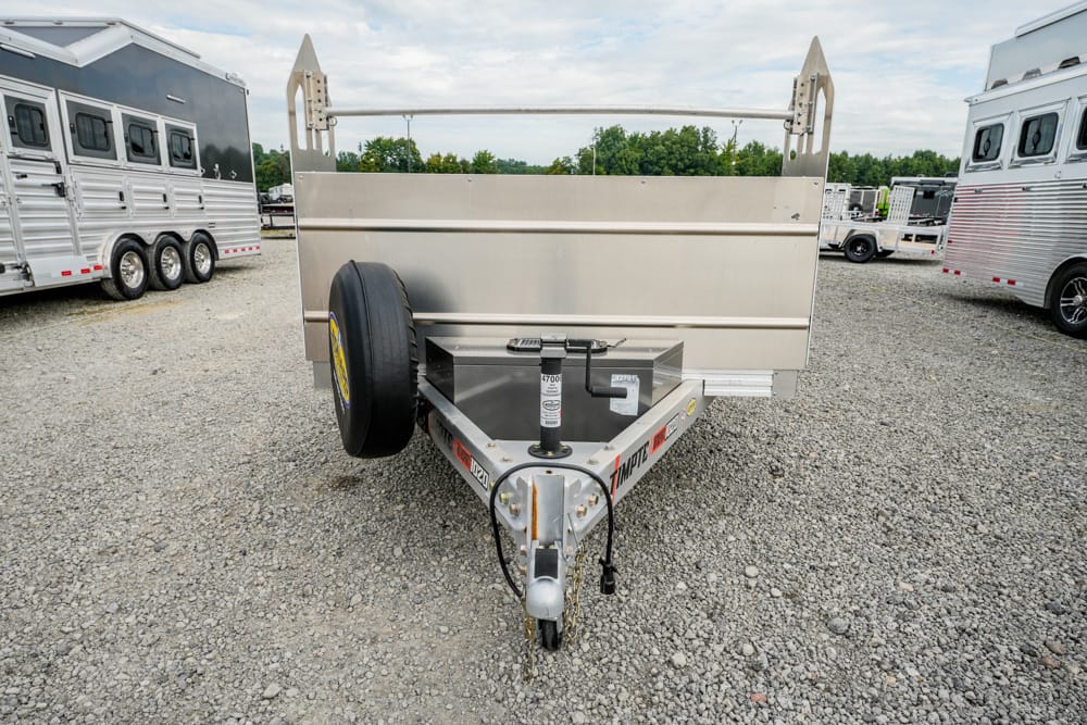 Front view of a silver utility trailer with a raised bed, spare tire on the left, and a hitch in a gravel lot with other trailers nearby.