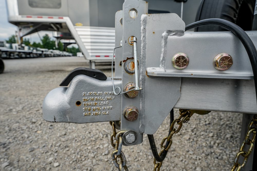 Close-up of a metal trailer hitch with bolts and safety chains on a gravel lot. (Informative)