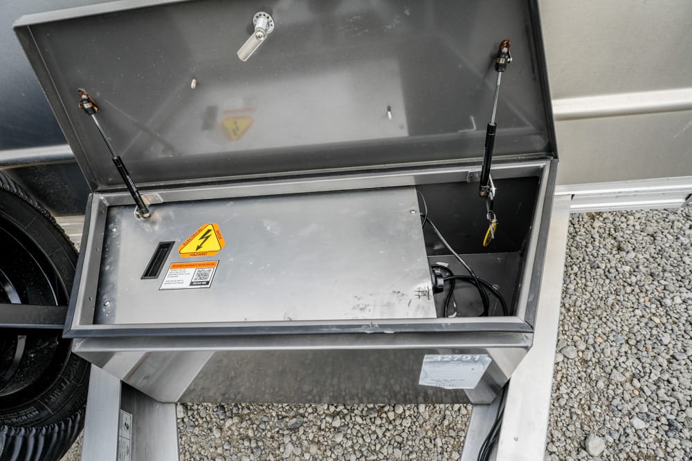 Open metal electrical cabinet with high-voltage warning sticker, exposing wiring inside a trailer setup on gravel surface.