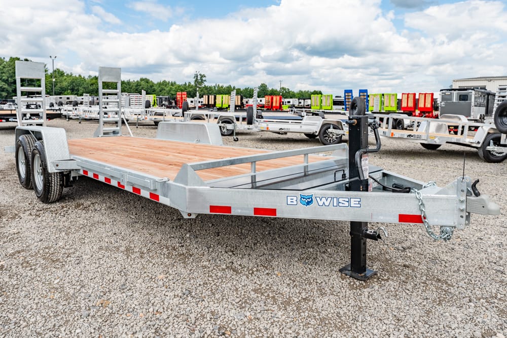 Flatbed trailer with a wooden deck and dual axles parked on a gravel lot, brand B-WISE visible on the frame.