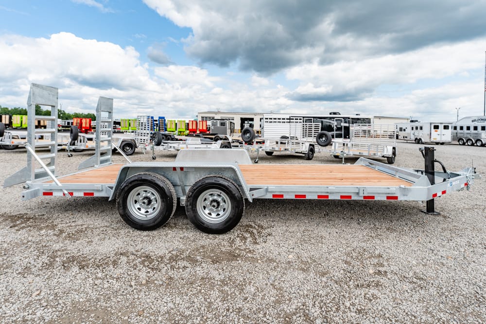 Double-axle flatbed trailer with a wooden deck and rear ramps in a gravel lot with other trailers nearby.